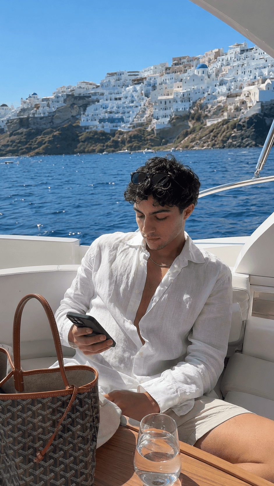 Man in a white linen shirt seated on a yacht deck, phone in hand, with the white cliffside village of Santorini across the water behind him